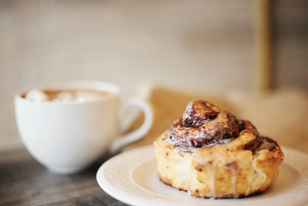 A steaming latte served in a white cup alongside a freshly baked cinnamon roll on a wooden table.