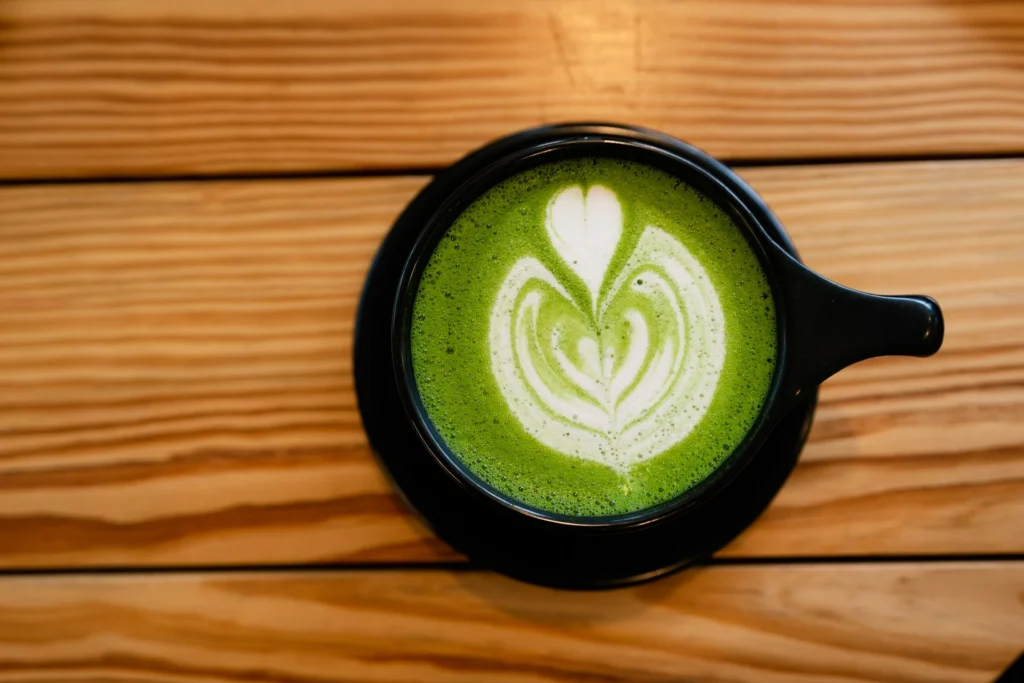 A cup of vibrant green matcha tea with a bamboo whisk and matcha powder on a wooden tray.